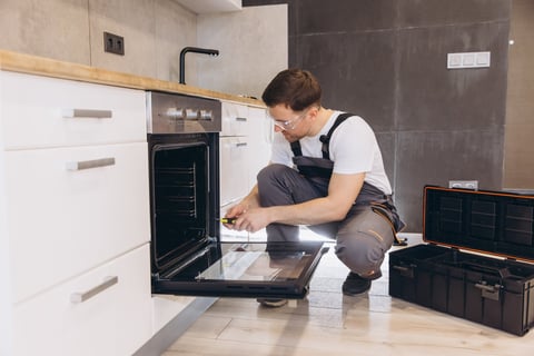Plumber wearing safety glasses using a screwdriver to repair an electric oven in a modern kitchen