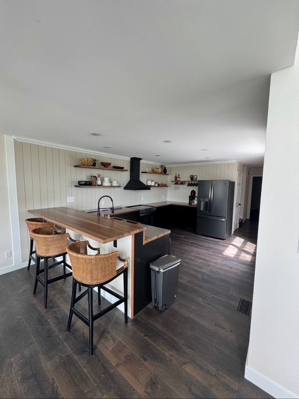 Modern kitchen with black island, woven bar stools, stainless steel appliances, open shelving, and dark wood flooring