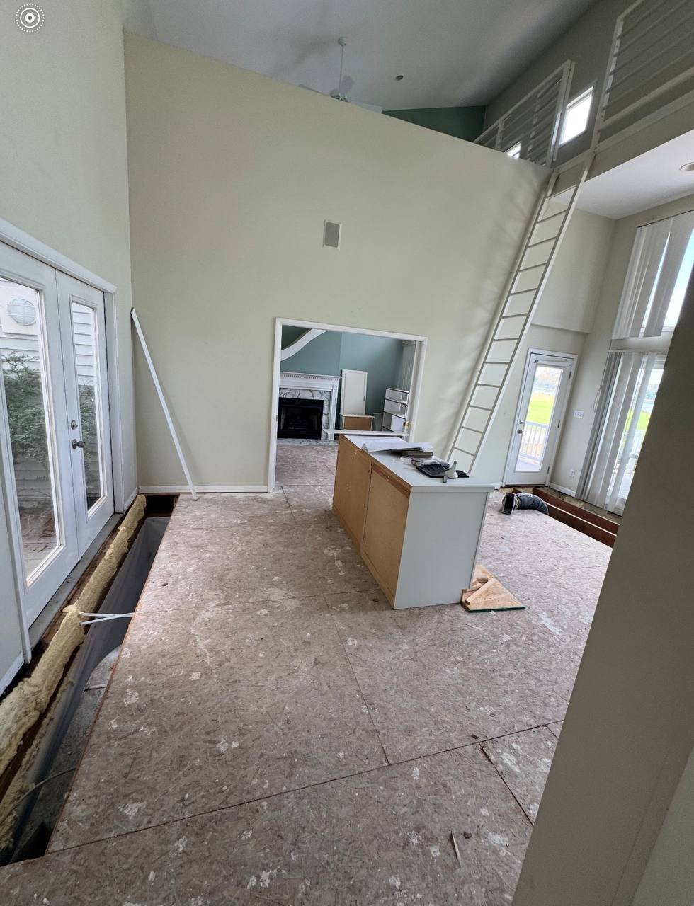 Interior of a home under renovation with exposed concrete floor, white walls, ladder, and kitchen counter visible in background